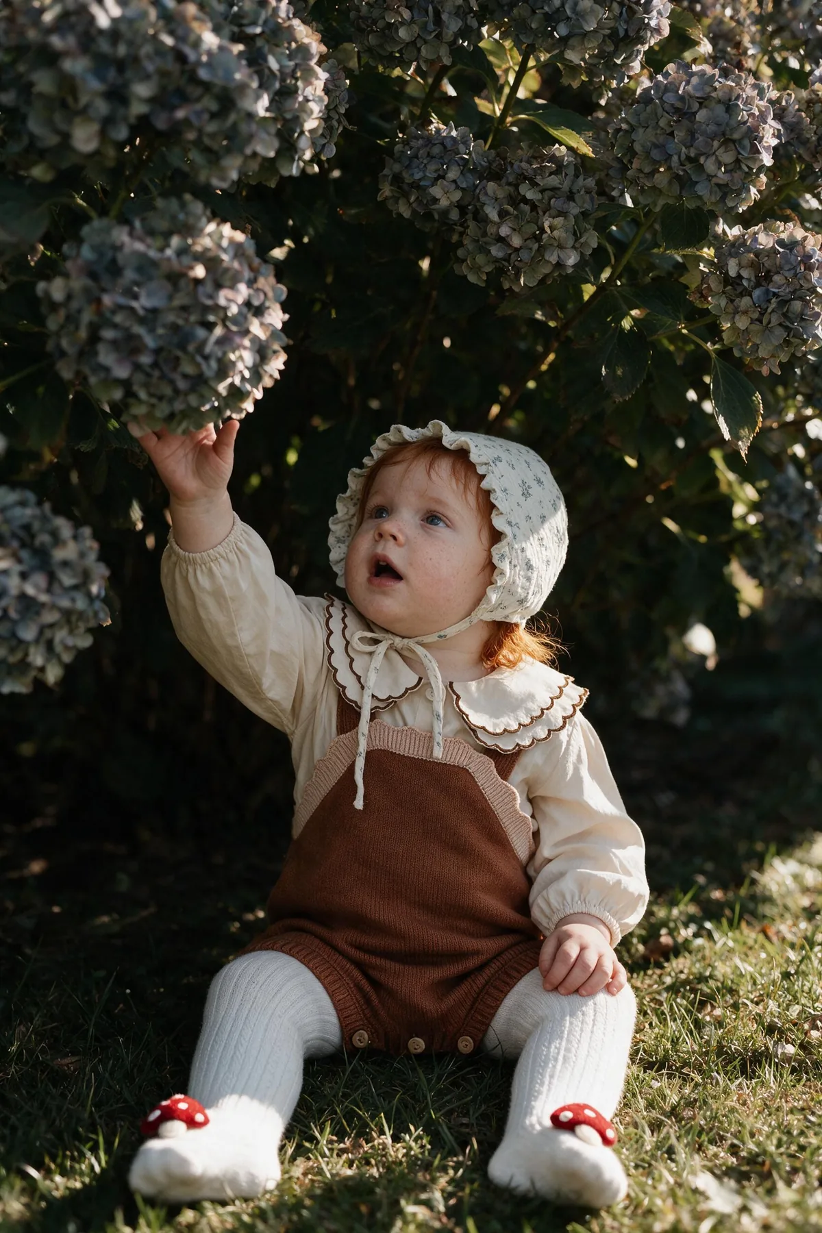 Baby girl in a rust knit romper, scallop-collar blouse, floral bonnet and toadstool tights reaching up to a faded hydrangea